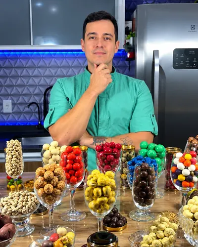 Gustavo Ono na cozinha atrás de uma mesa cheia de taças e potes de drágeas e trufas coloridas