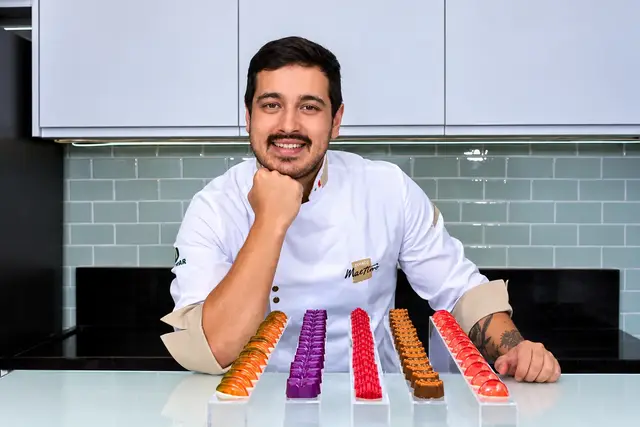 Gustavo Ono, de dólmã branca, sorrindo com a mão no queixo atrás de fileiras de bombons coloridos